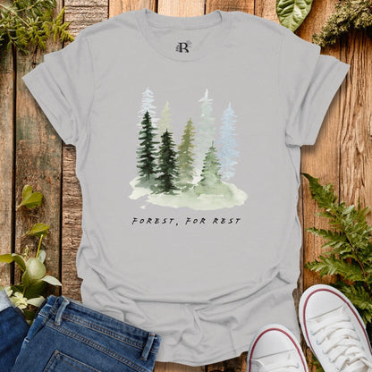 Gray t-shirt with forest design and 'Forest, For Rest' text on a wooden background with plants.