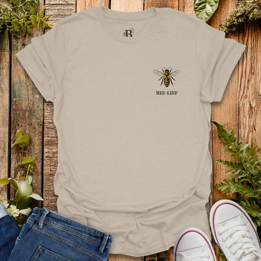 Beige t-shirt with a bee design and 'BEE-LIEF' text on a wooden background with plants.