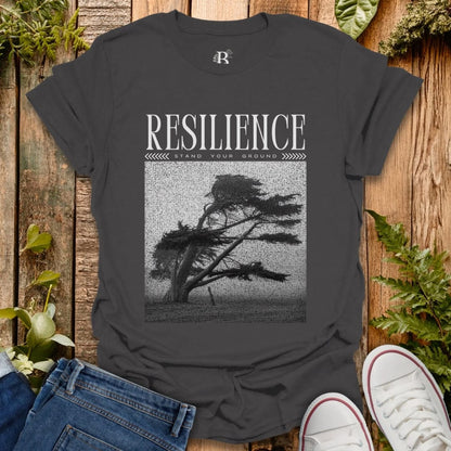 Dark gray t-shirt with 'Resilience' graphic design on a wooden surface with plants.