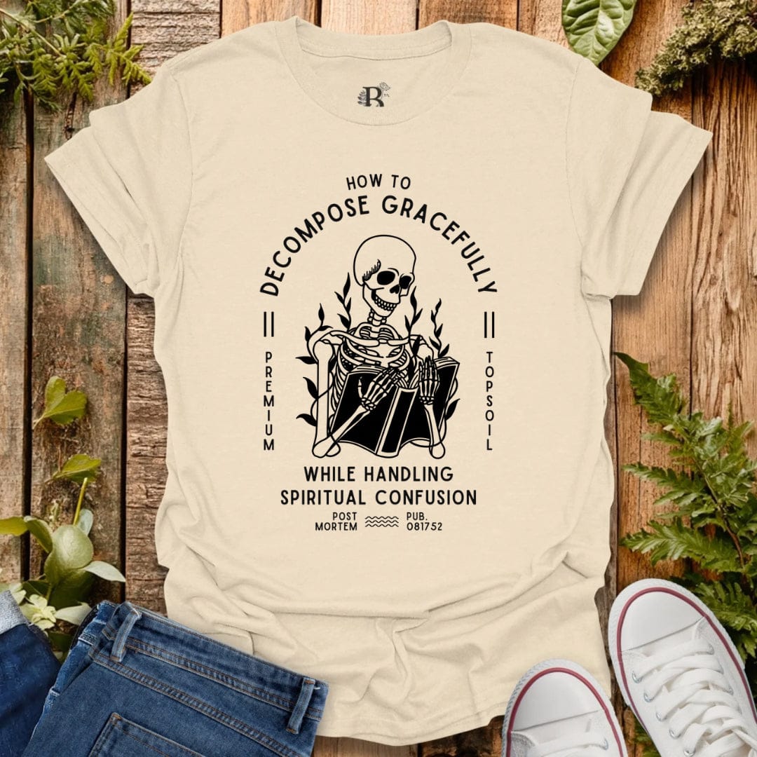 Beige t-shirt with a graphic design featuring a skeleton and text, displayed on a wooden surface with plants.