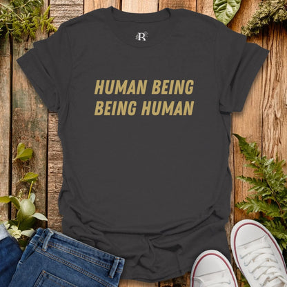 Black t-shirt with 'HUMAN BEING BEING HUMAN' text on a wooden background with jeans and sneakers.