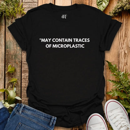 Black t-shirt with text 'MAY CONTAIN TRACES OF MICROPLASTIC' on a wooden surface with plants.