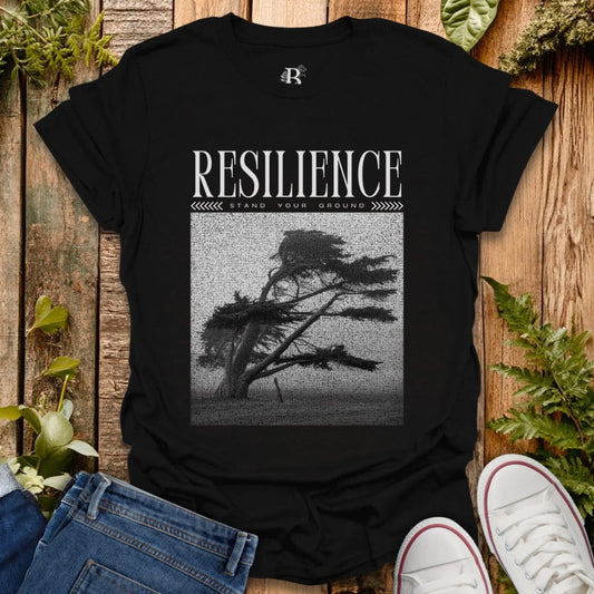 Black t-shirt with 'Resilience' graphic and text on a wooden surface with plants.