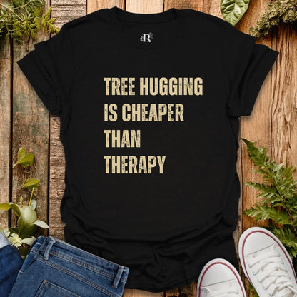 Black t-shirt with 'Tree Hugging is Cheaper Than Therapy' text on a wooden background with plants.