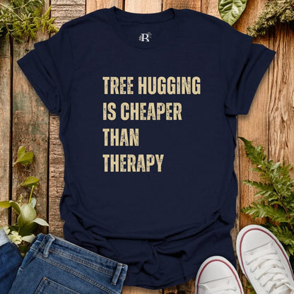 Navy blue t-shirt with 'Tree Hugging is Cheaper Than Therapy' text on a wooden background with plants.
