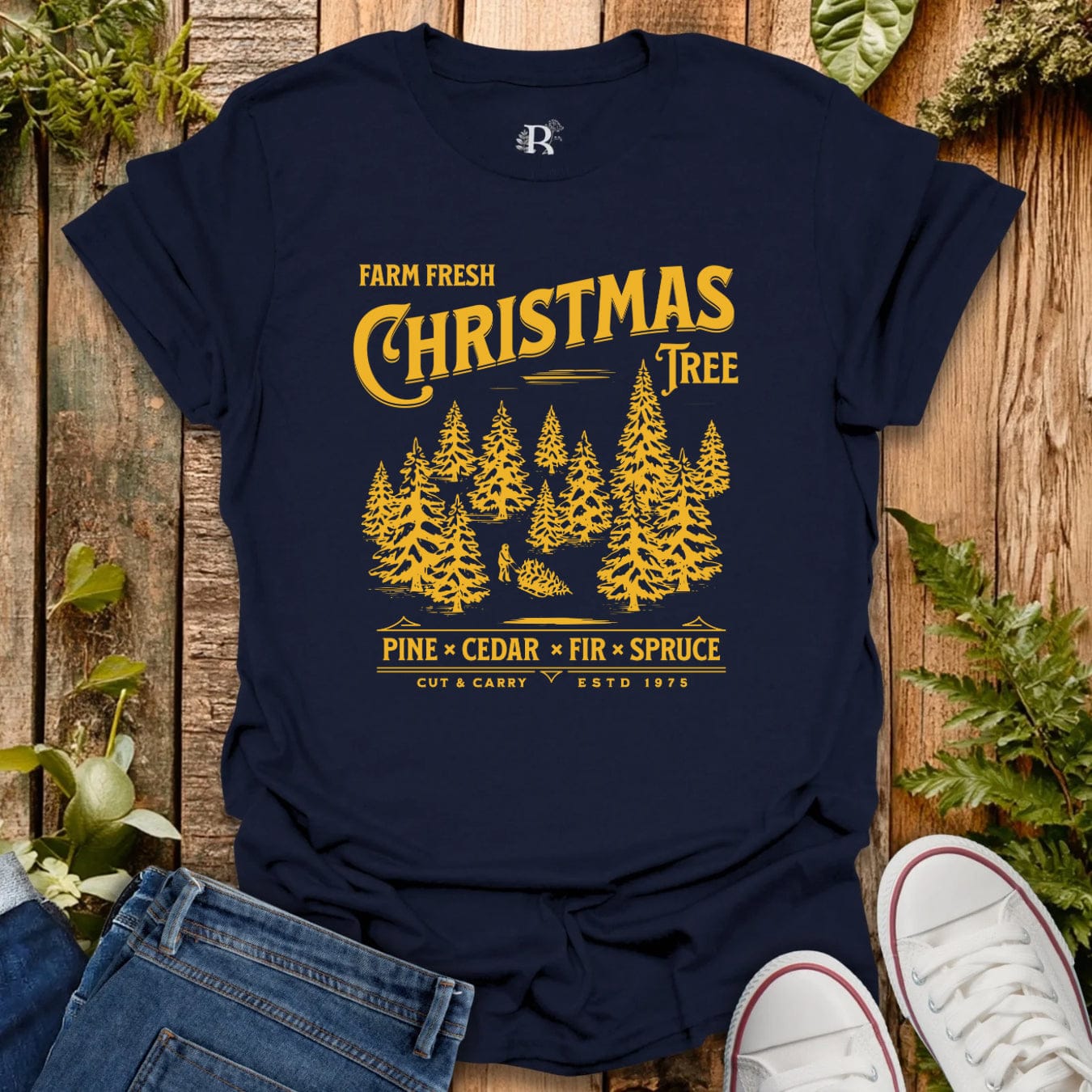 Navy blue t-shirt with yellow Christmas tree design on a wooden surface with greenery.
