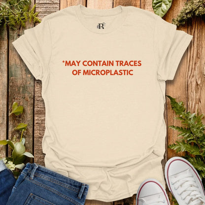 Beige t-shirt with red text 'MAY CONTAIN TRACES OF MICROPLASTIC' on a wooden background with plants.