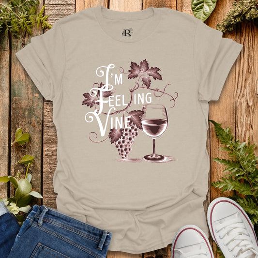 Beige t-shirt with 'I'm Feeling Vine' graphic design on a wooden background with plants.