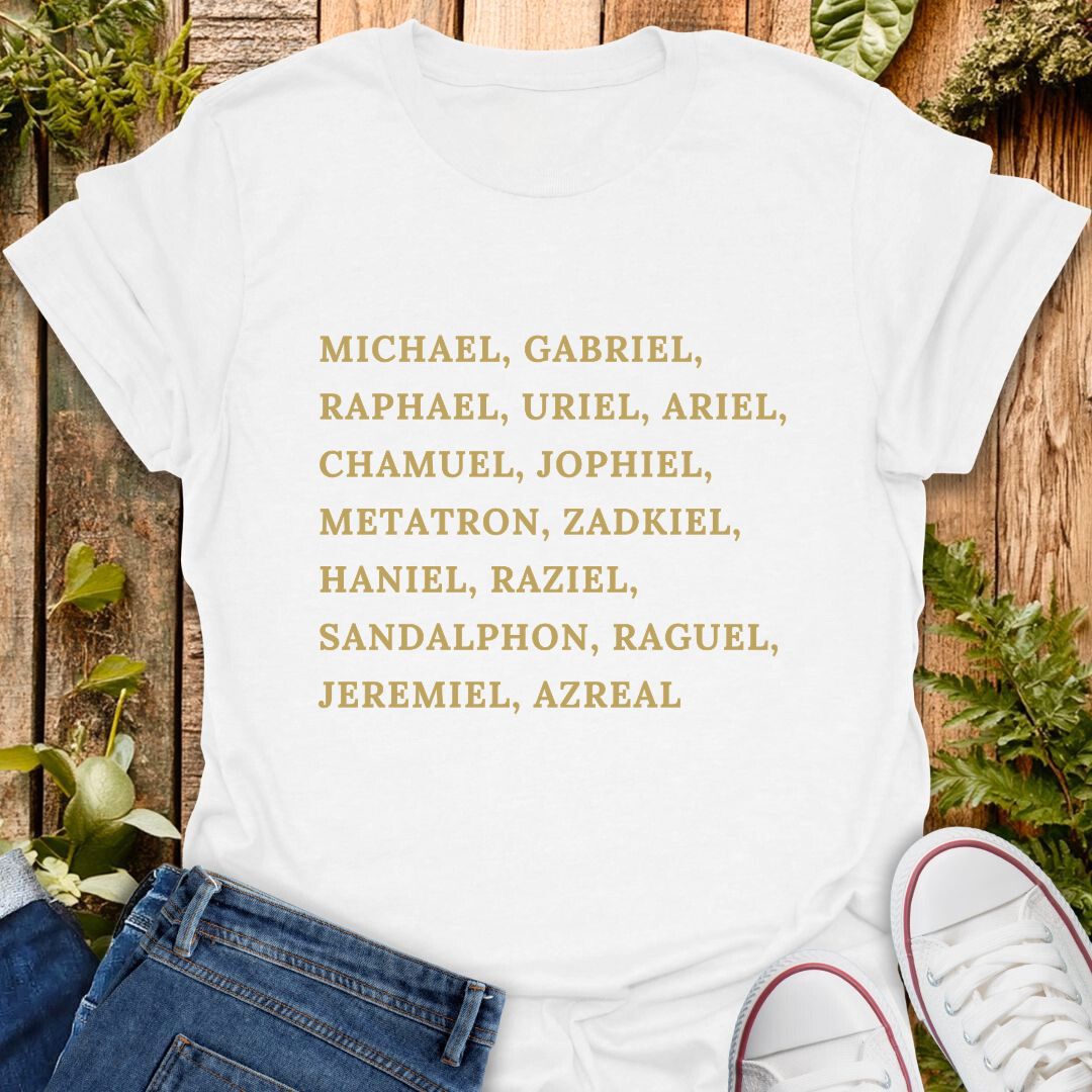 A white short-sleeve T-shirt laid flat on a rustic wooden surface with greenery and jeans, with a pair of white sneakers partly visible at the bottom right. The shirt has a list of names of archangels printed in gold text of on the front.
