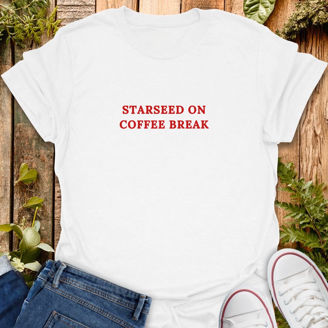 White t-shirt with red text 'Starseed on coffee break' text on a wooden surface with plants.